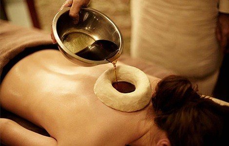 Warm herbal oil being poured into a dough ring placed on a client's upper back